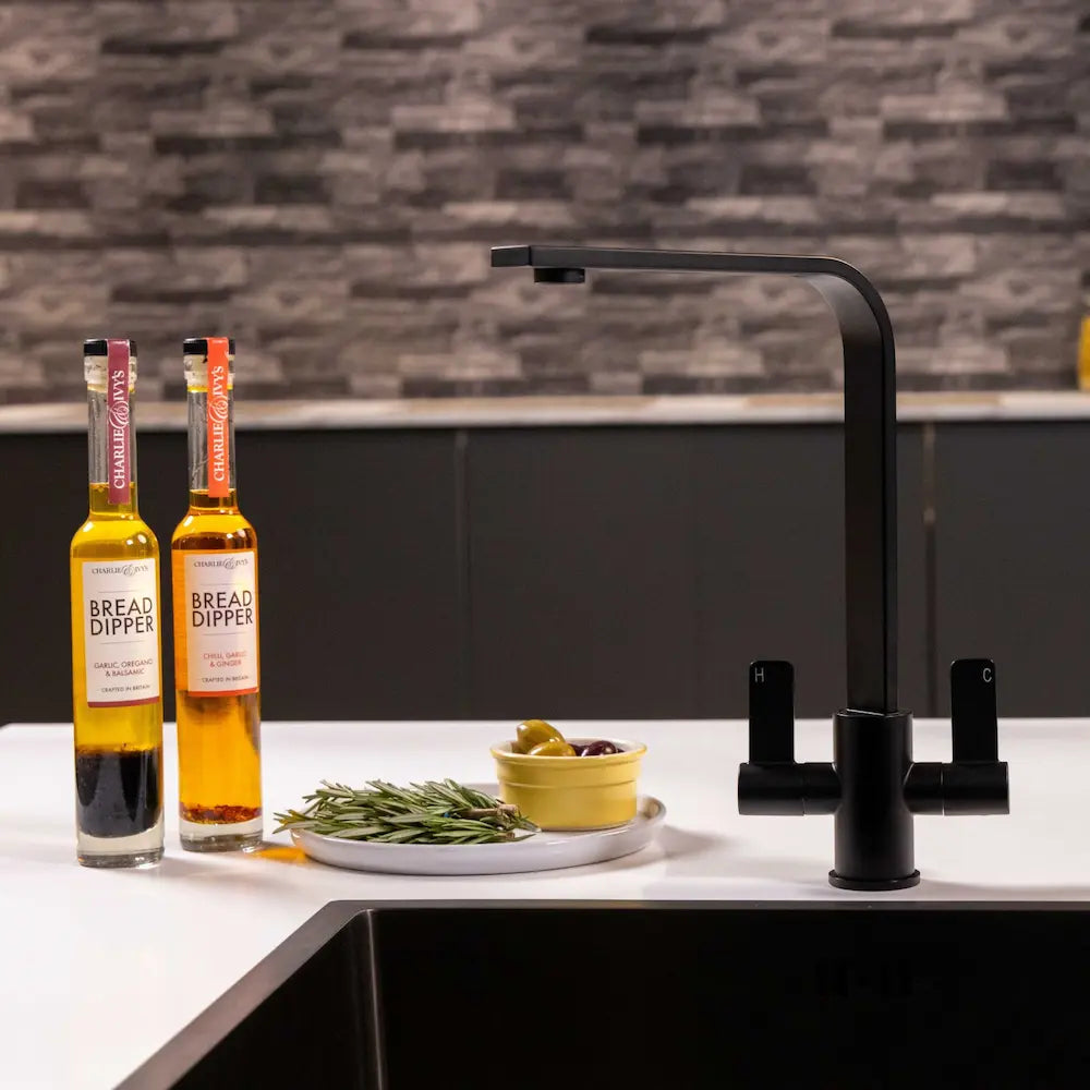 Matt black twin lever kitchen tap above a black sink with a brick splashback behind and oil bottles on the worktop