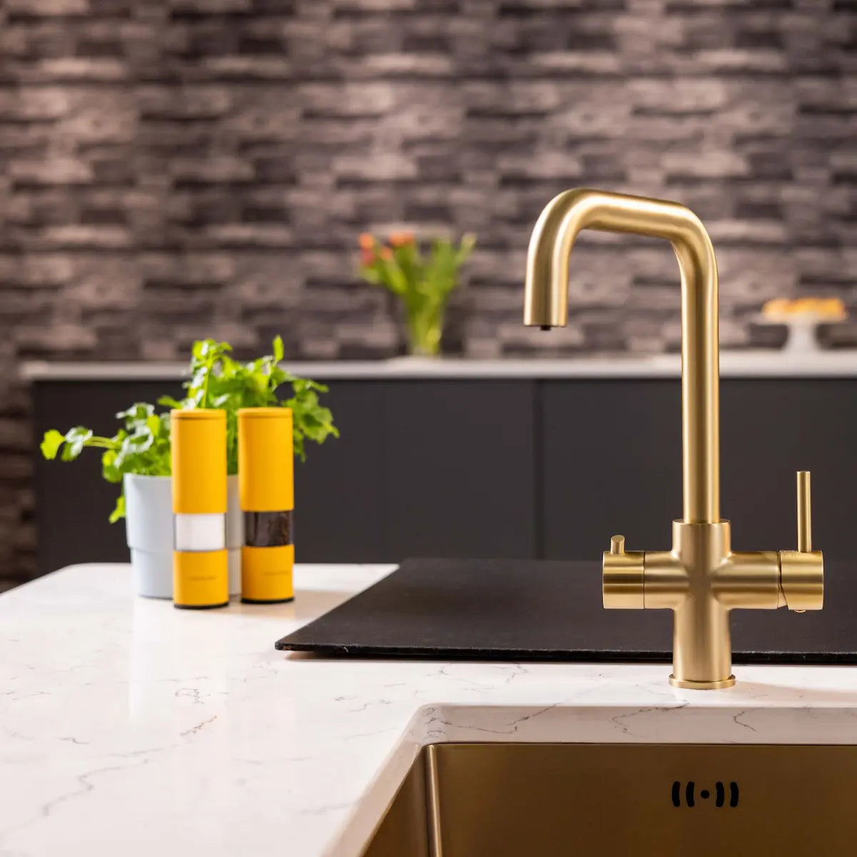 Brushed brass 3 in 1 boiling tap on a white marble kitchen island with a brass sink, set against a grey brick splashback
