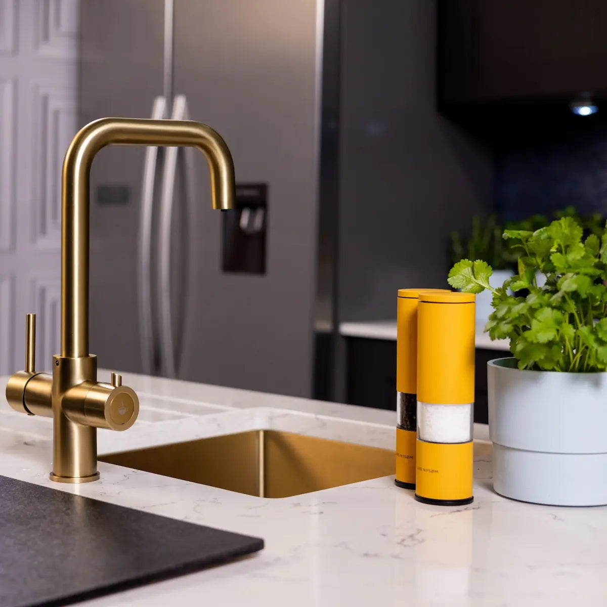 Brushed brass boiling tap seen at an angle on a marble worktop beside a brass sink, with a stainless steel fridge in the background