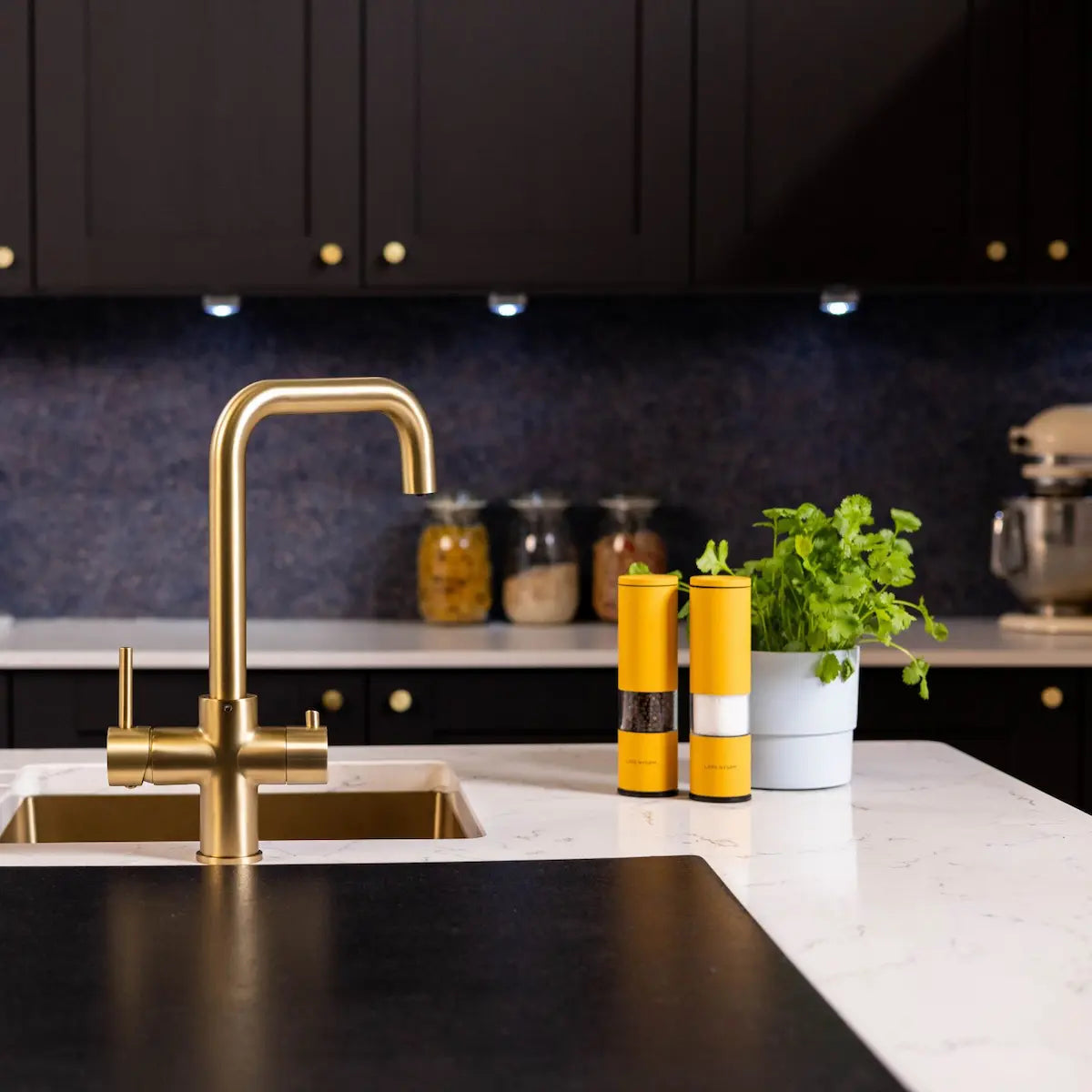 Brushed brass boiling water kitchen tap on a white marble island above a brass undermount sink, with navy splashback and black cabinets behind