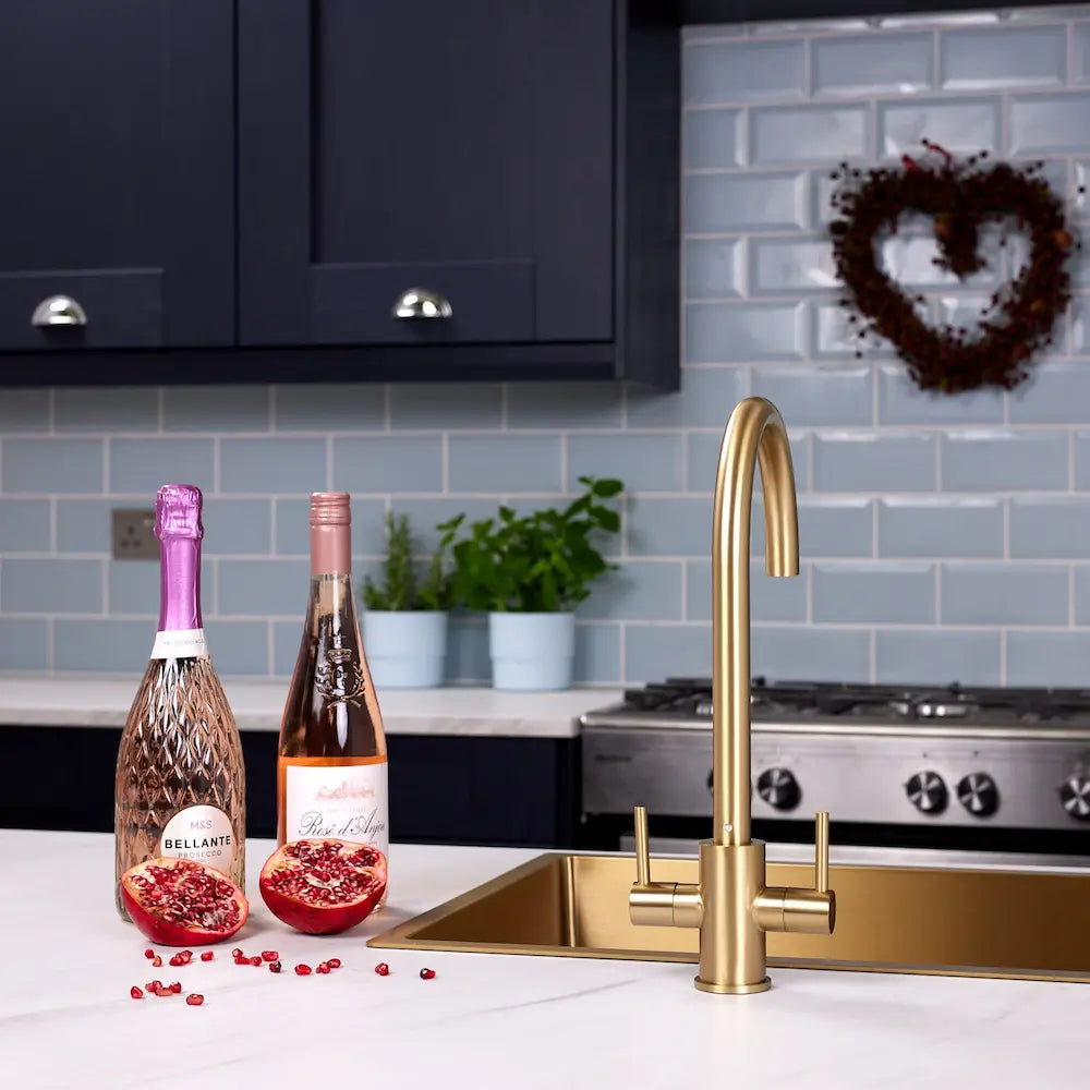 Brushed brass twin lever kitchen tap on a white worktop beside a range cooker in a navy kitchen