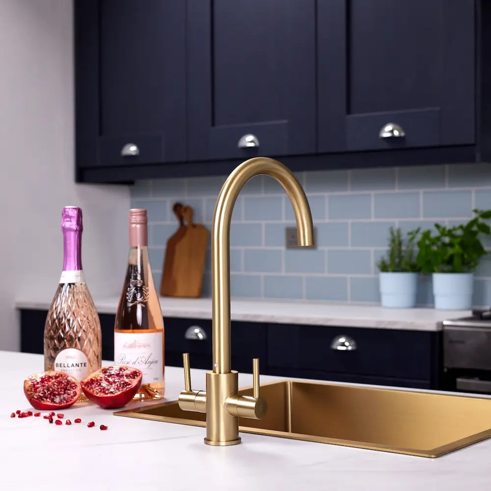 Brushed brass dual lever kitchen tap over a brass sink, with navy shaker units and pale blue metro tiles behind