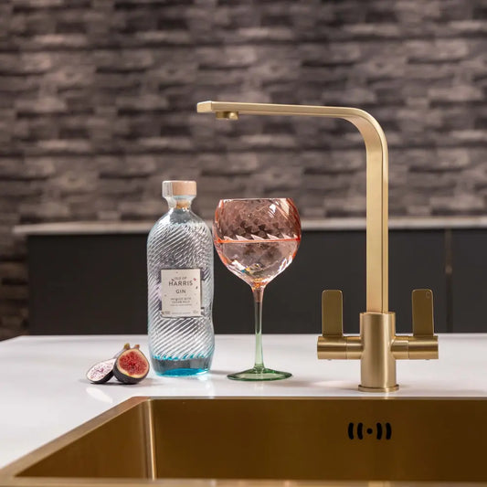 Brushed brass twin lever kitchen tap above a gold undermount sink on a white worktop, with a brick splashback behind