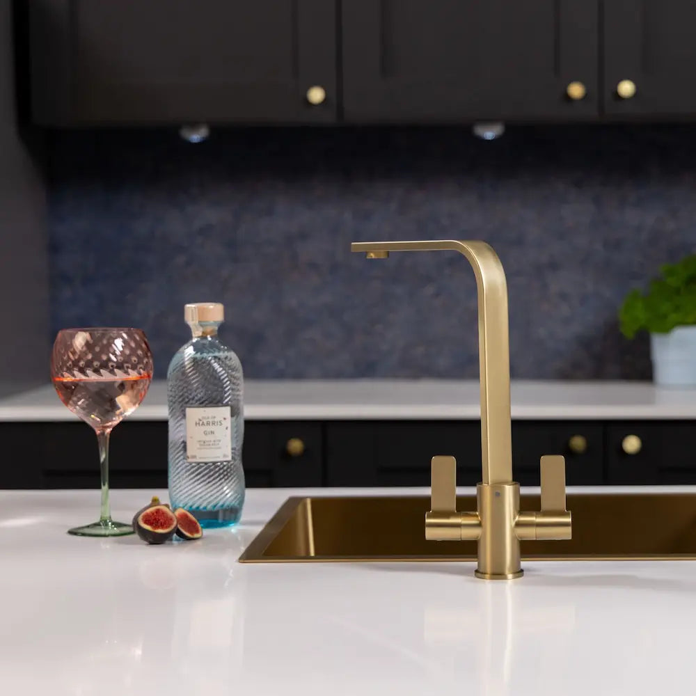 Brushed brass twin lever tap beside a gold sink in a grey panelled kitchen