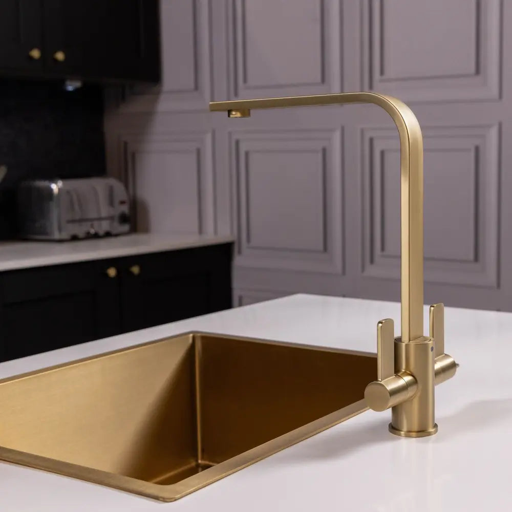 Brushed brass kitchen tap fitted to a white worktop in a dark navy kitchen, viewed across the room