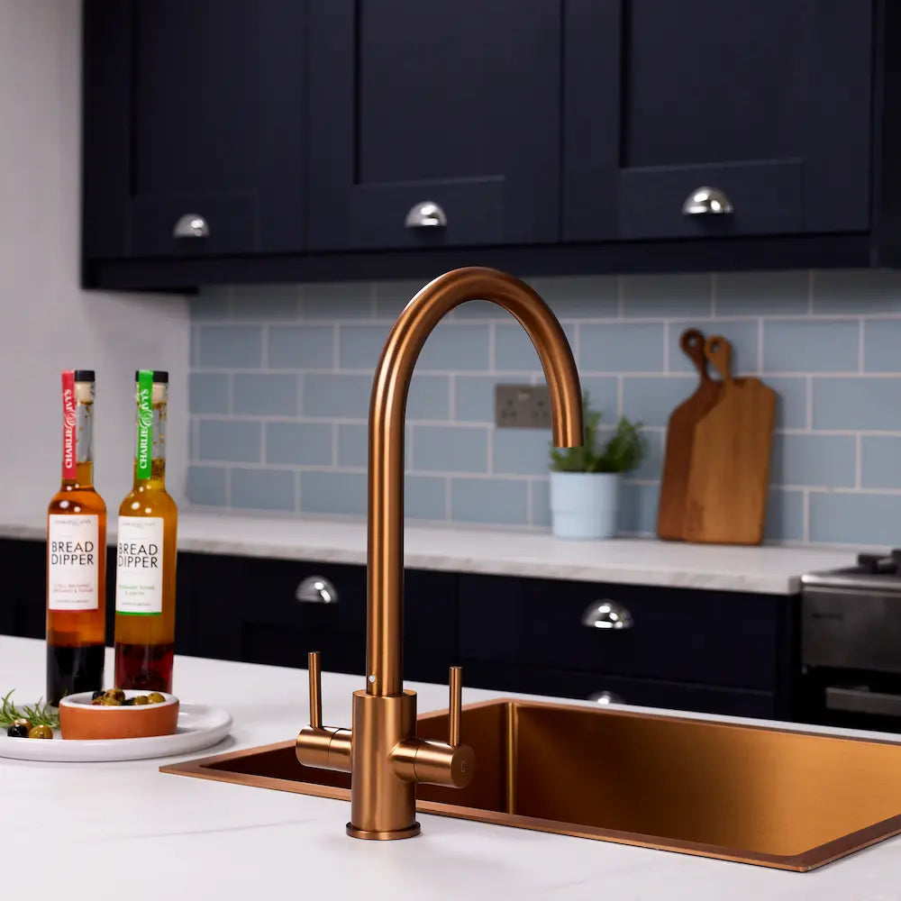 Adria brushed copper kitchen tap with a tall swan neck spout above a copper sink, navy units and herbs behind