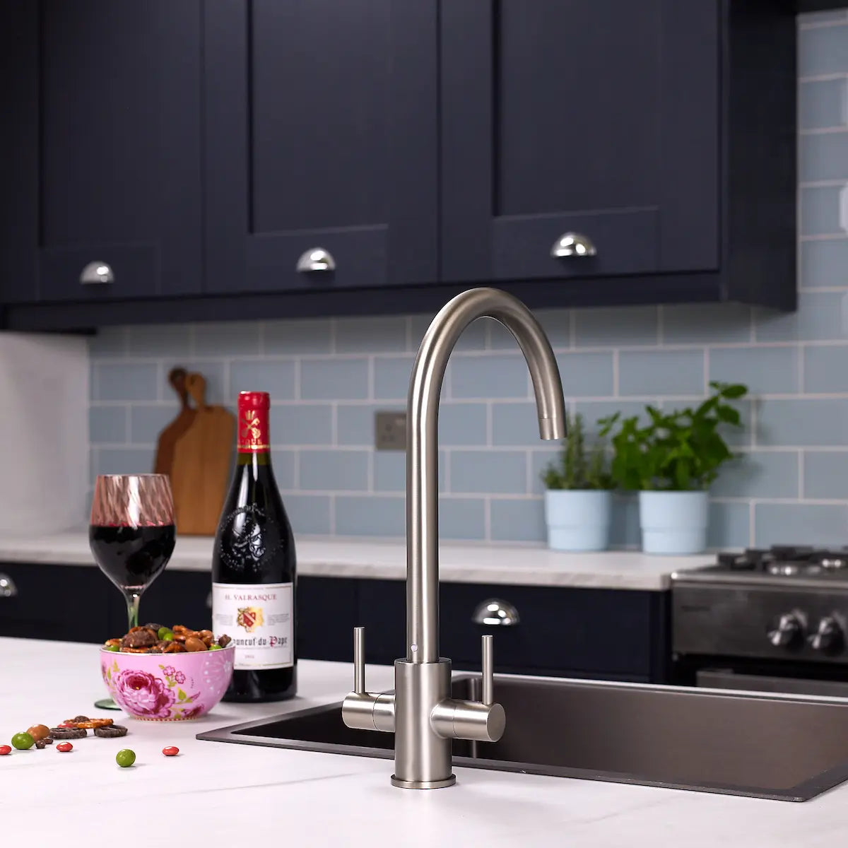 Brushed steel kitchen tap above a stainless sink in a navy kitchen with a gas range and blue tiled splashback