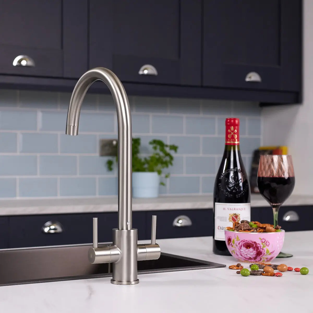 Brushed steel dual lever kitchen tap fitted beside a stainless steel sink in a navy shaker kitchen with blue metro tiles
