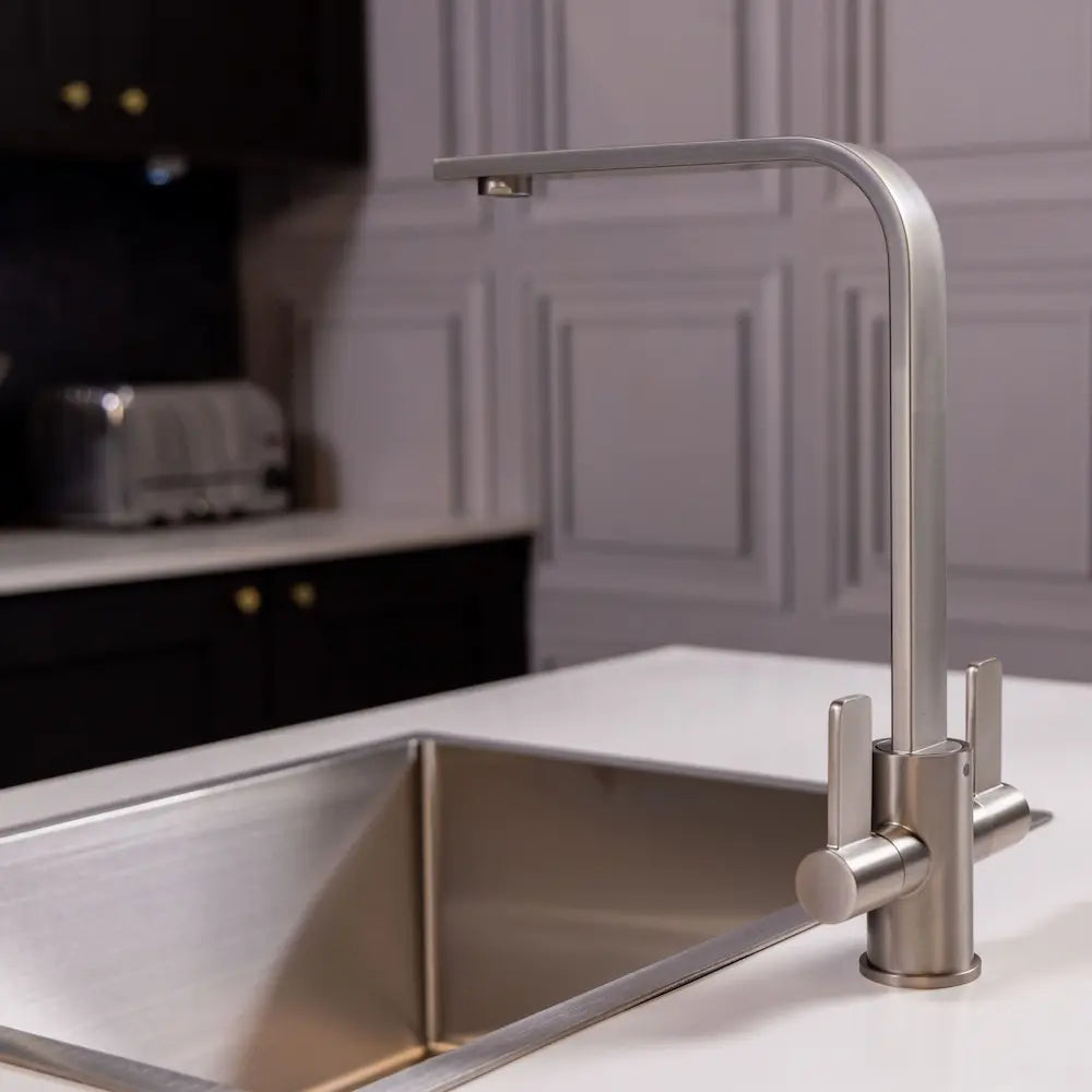 Side view of the brushed steel twin lever tap beside a stainless sink, with black units and a toaster in the background