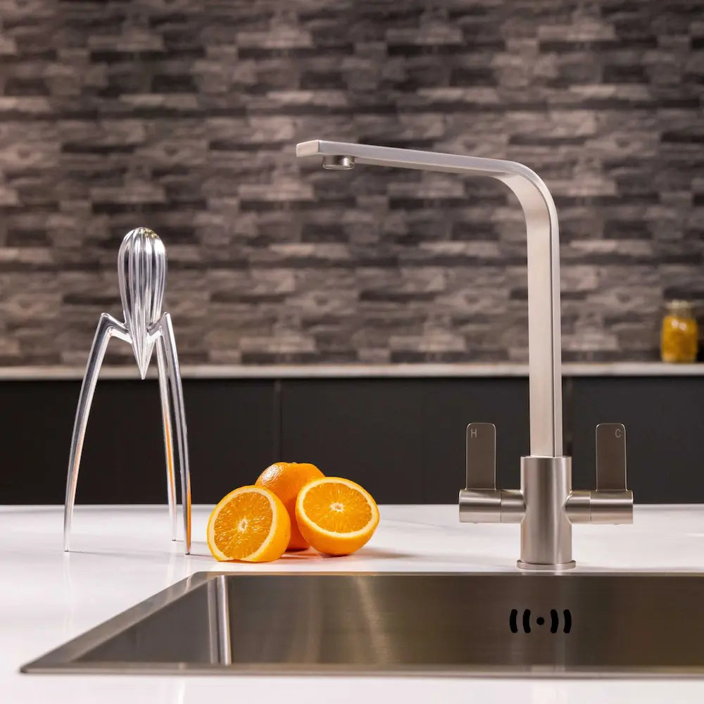 Brushed steel twin lever kitchen tap on a white worktop against a grey stone-effect splashback, with oranges and a citrus squeezer beside it