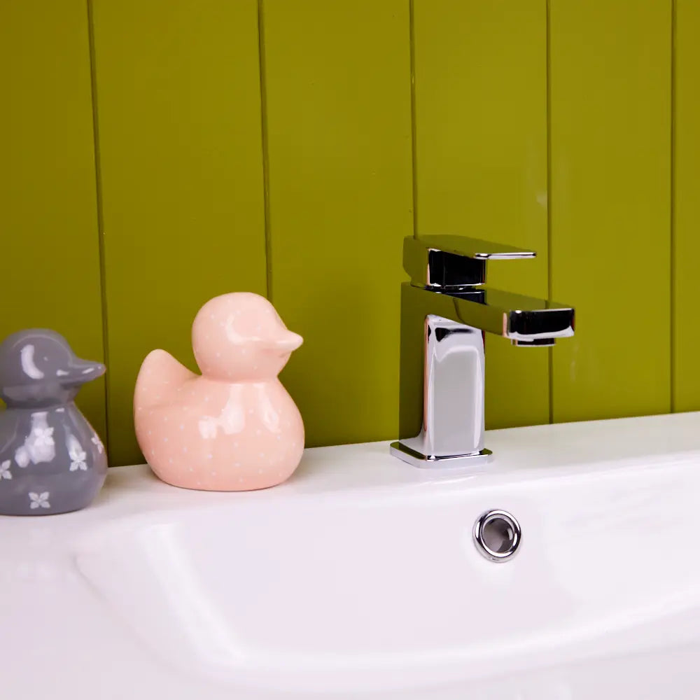Chrome Lanza basin tap fitted to a white basin against an olive green panelled wall, with the chrome waste visible in the bowl below.