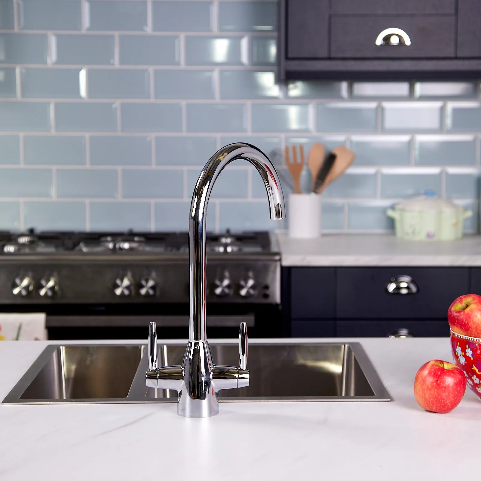 Chrome dual lever kitchen tap fitted to a stainless steel sink in a navy kitchen, with pale blue metro tiles and a range cooker behind