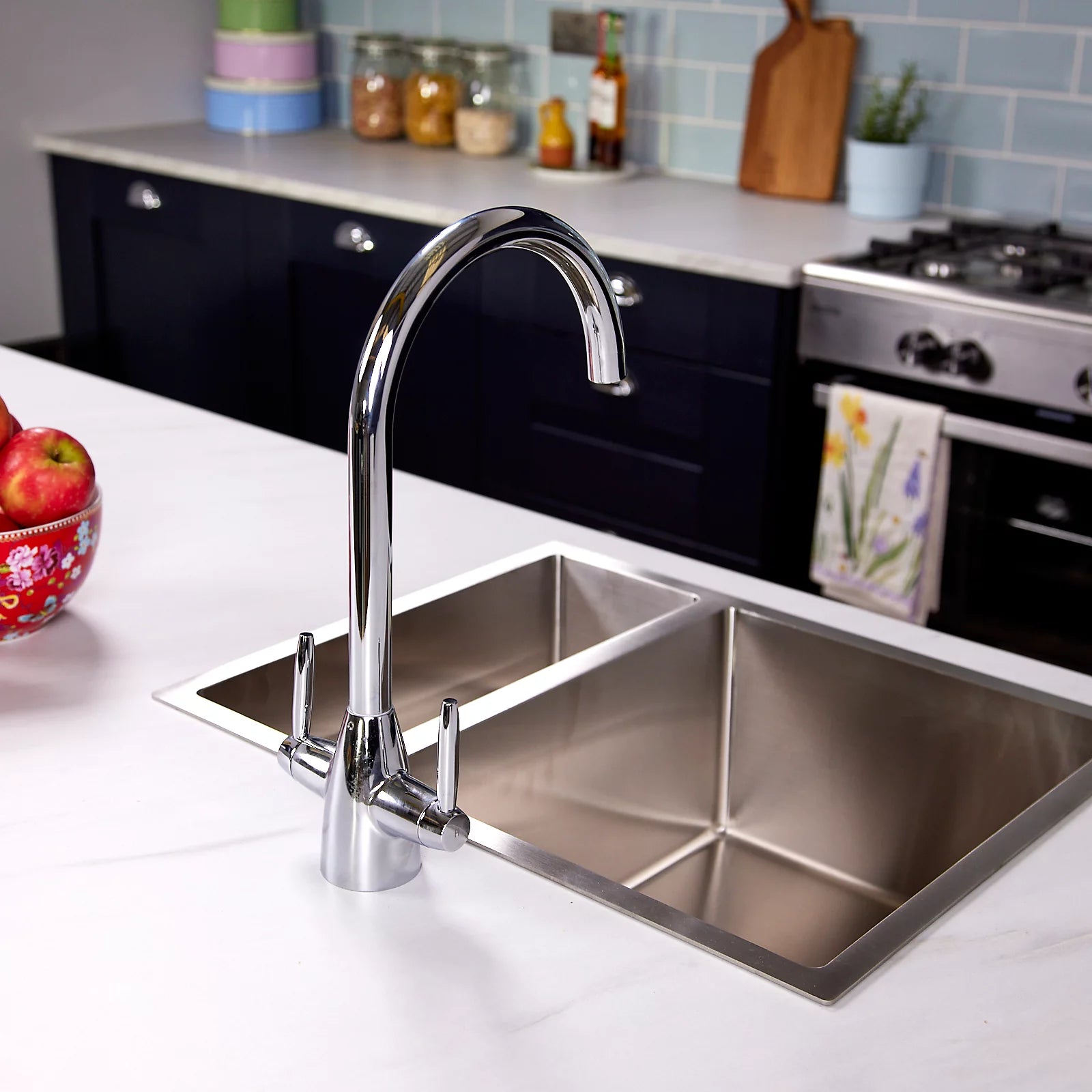 Chrome Astrid kitchen tap beside a large stainless steel sink, with navy cabinets and storage jars on the shelf behind