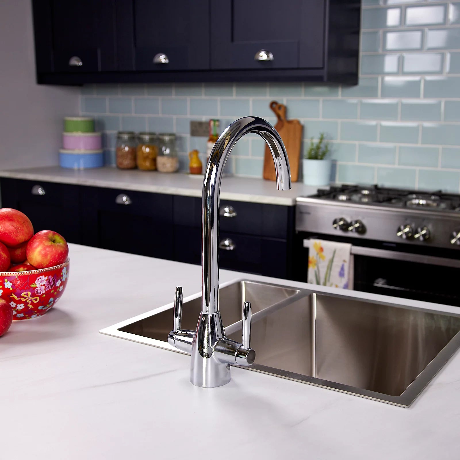 Chrome dual lever kitchen tap on a white worktop viewed from the side, with a bowl of apples beside the stainless sink