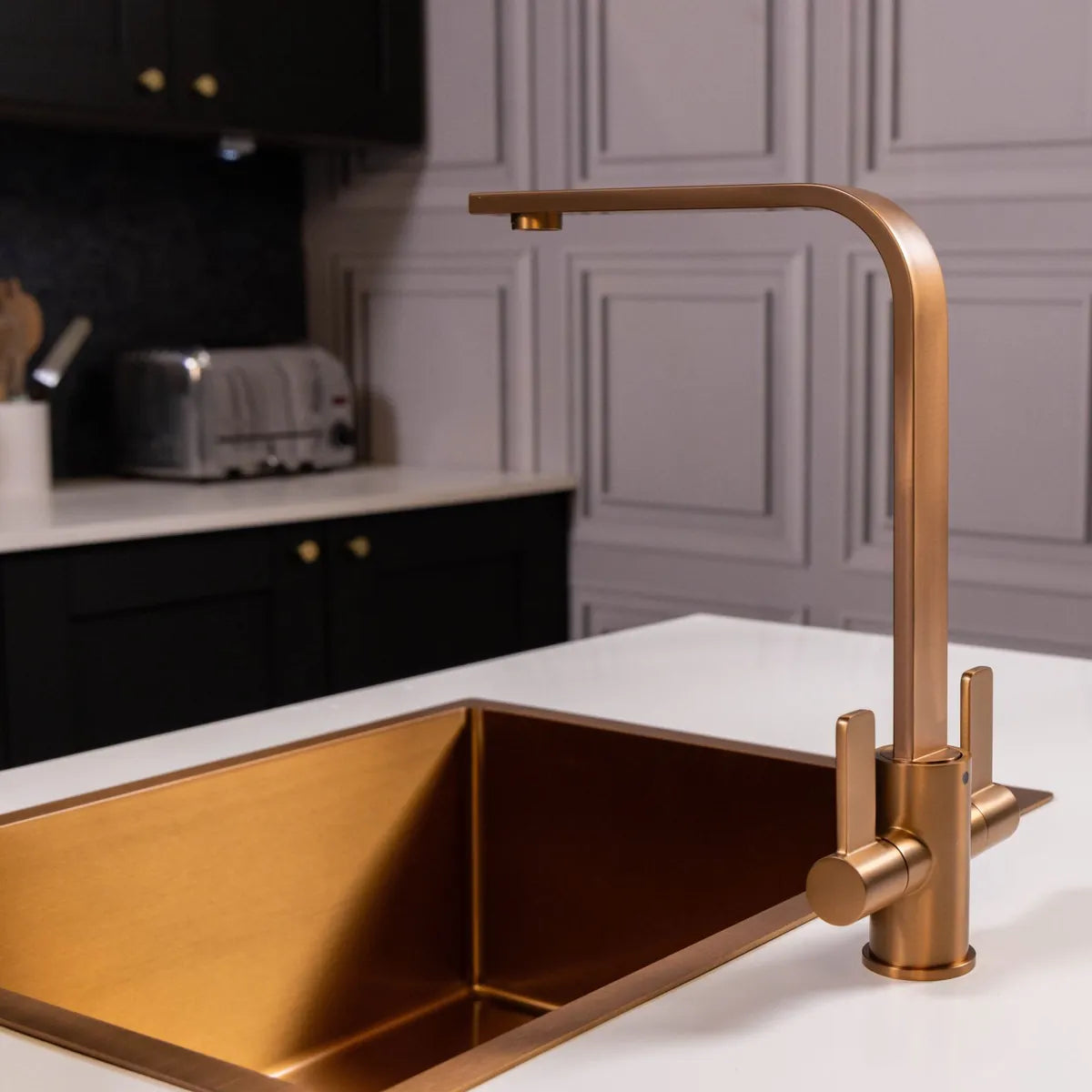 Copper kitchen mixer tap beside a copper-coloured sink in a grey panelled kitchen, with a toaster on the worktop behind