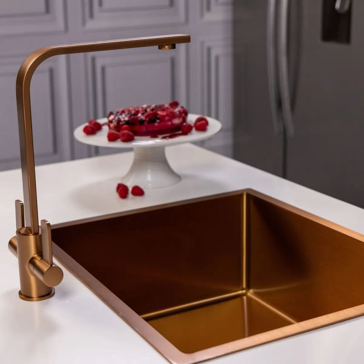 Side view of the brushed copper twin lever tap over a copper-coloured sink, with a raspberry cake on a stand behind