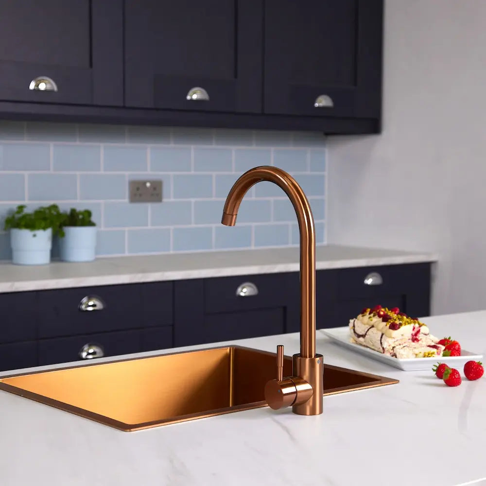 Copper swan neck kitchen mixer tap beside a brushed copper sink on a white marble-effect worktop