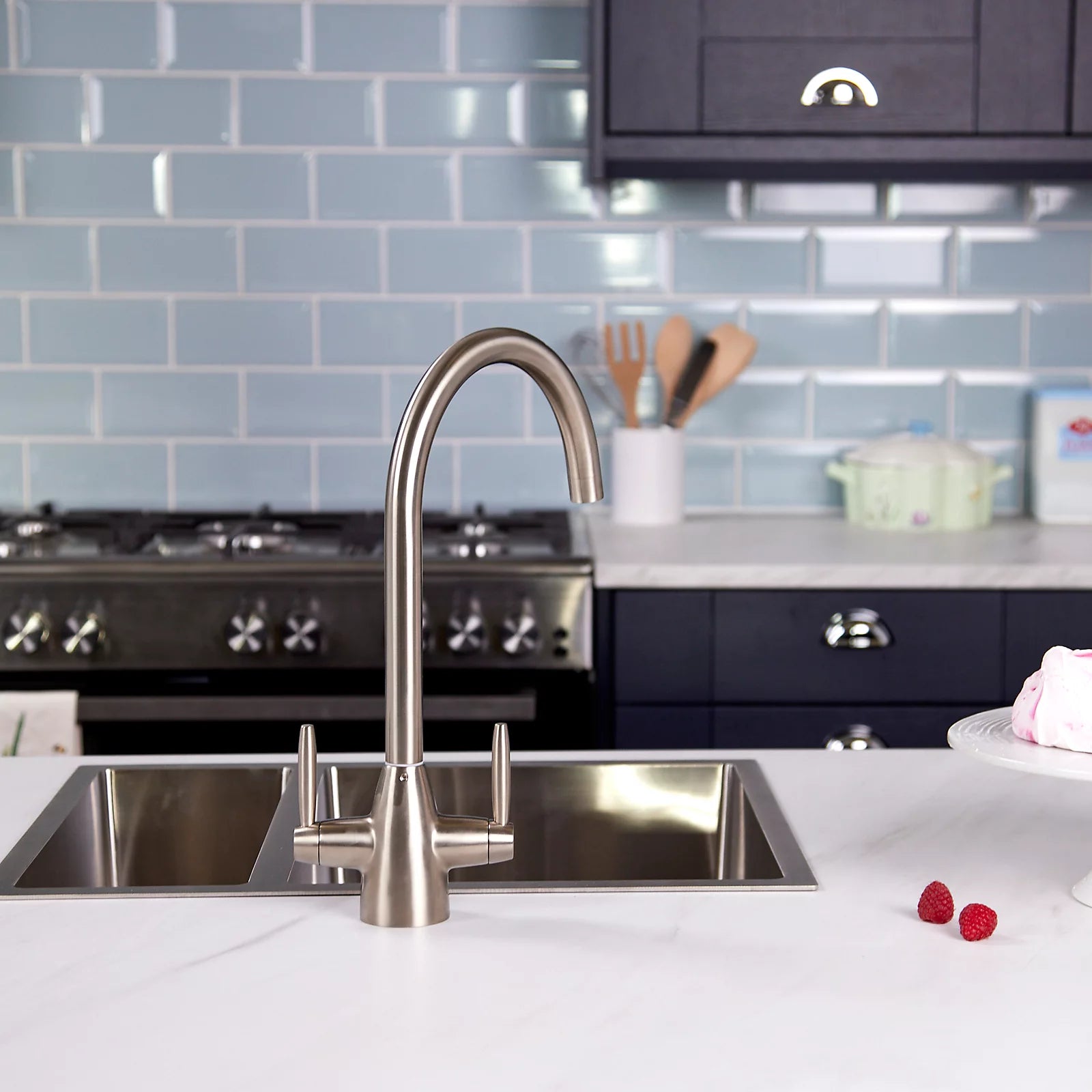 Pewter dual lever kitchen tap on a double stainless steel sink, with pale blue metro tiles and a range cooker behind