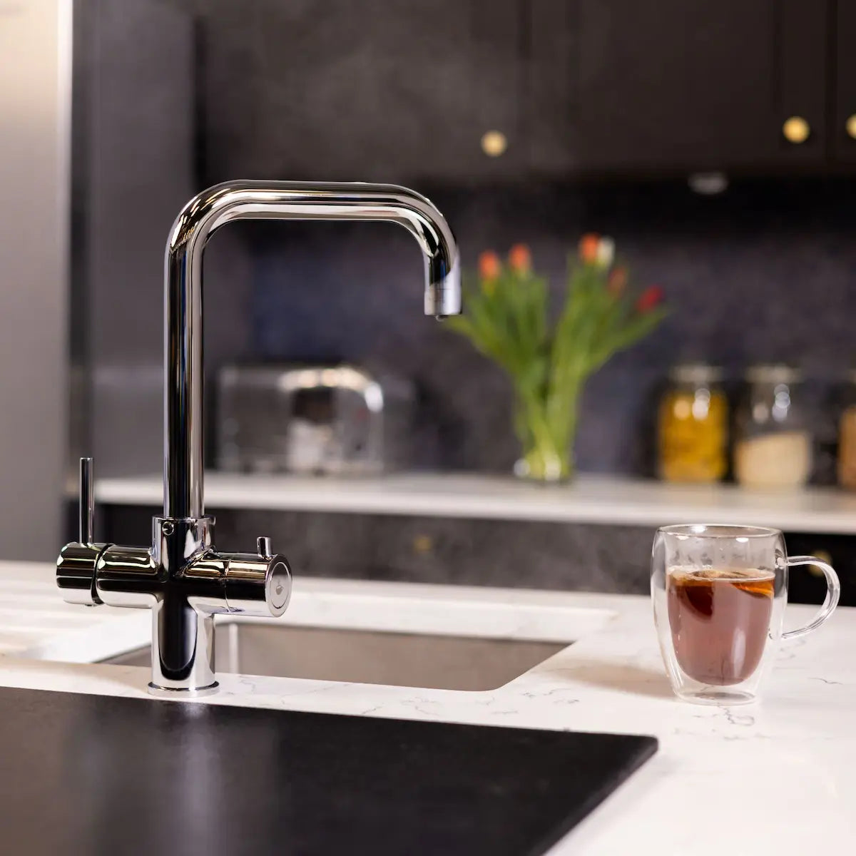 Chrome instant boiling water tap fitted to a white marble worktop above a stainless sink, with a glass of tea beside it in a dark kitchen