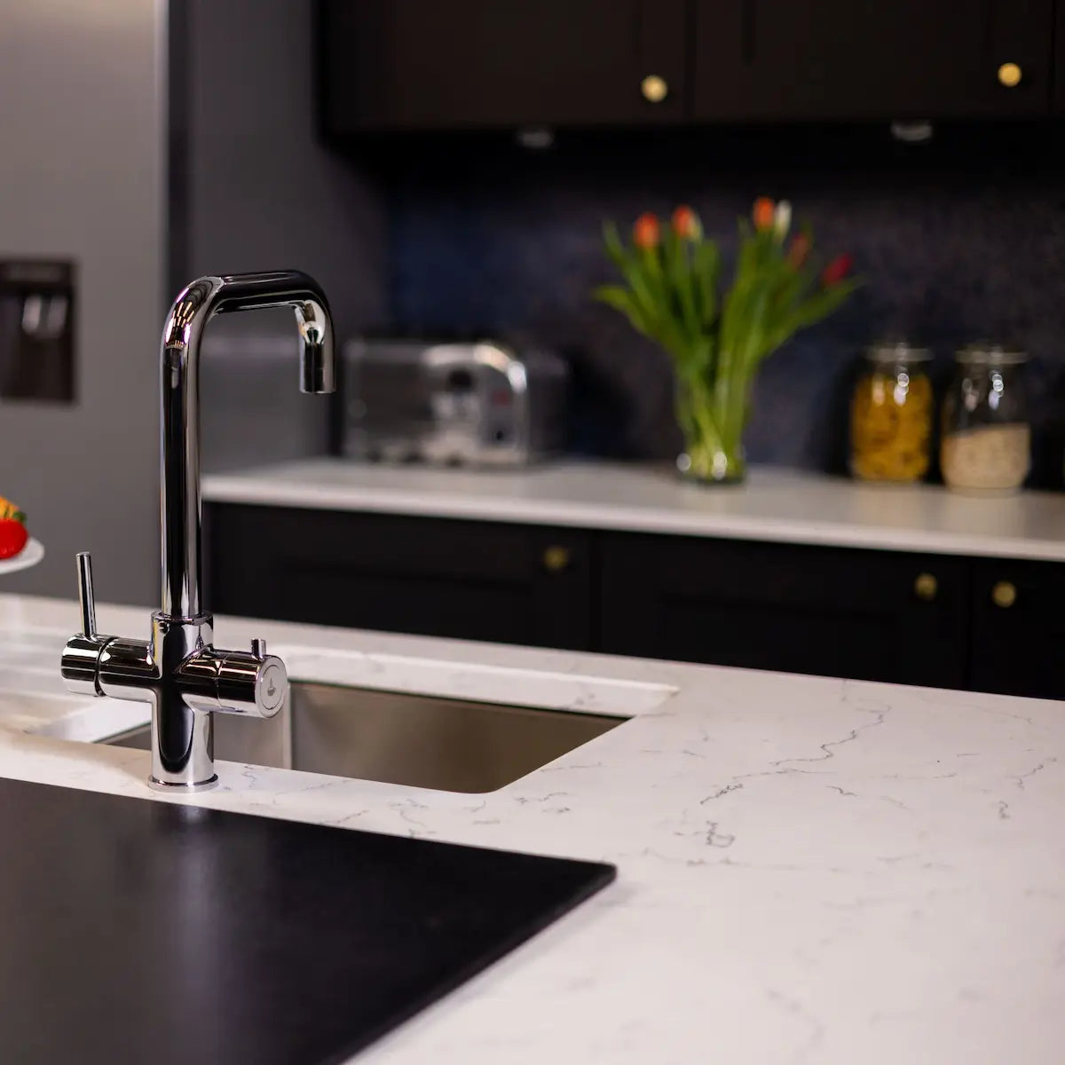 Wider view of the chrome boiling water tap on a marble worktop, with dark cabinets and a navy splashback behind
