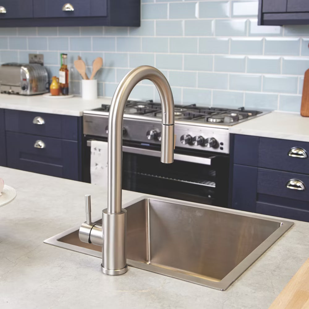The brushed steel Cato kitchen tap fitted to a stainless steel sink in a navy kitchen, with the spray head docked in the gooseneck spout.